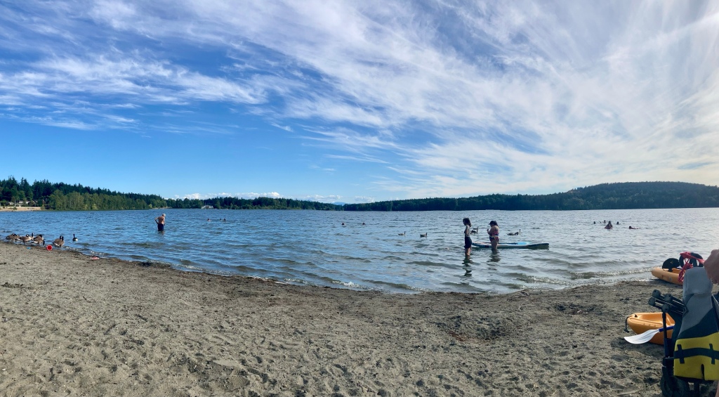 View of lake from beach on a sunny day. Someone in the water has a paddle board.